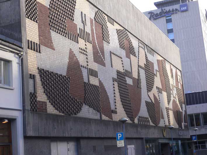 Fasaden til Agder Teater er et veggrelieff med brune, gule og grå murstein danner et geometrisk tema av mørke figurer mot lys grunn