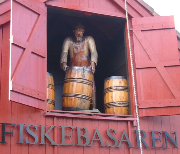 Helfigur i furu av mann med fisk som står i en takluke på fiskebrygga i Kristiansand
