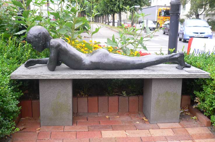 Liggende figur i støpt bronse og er plassert på en liggende marmorsokkel utenfor ”Snadderkiosken” i Elvegata